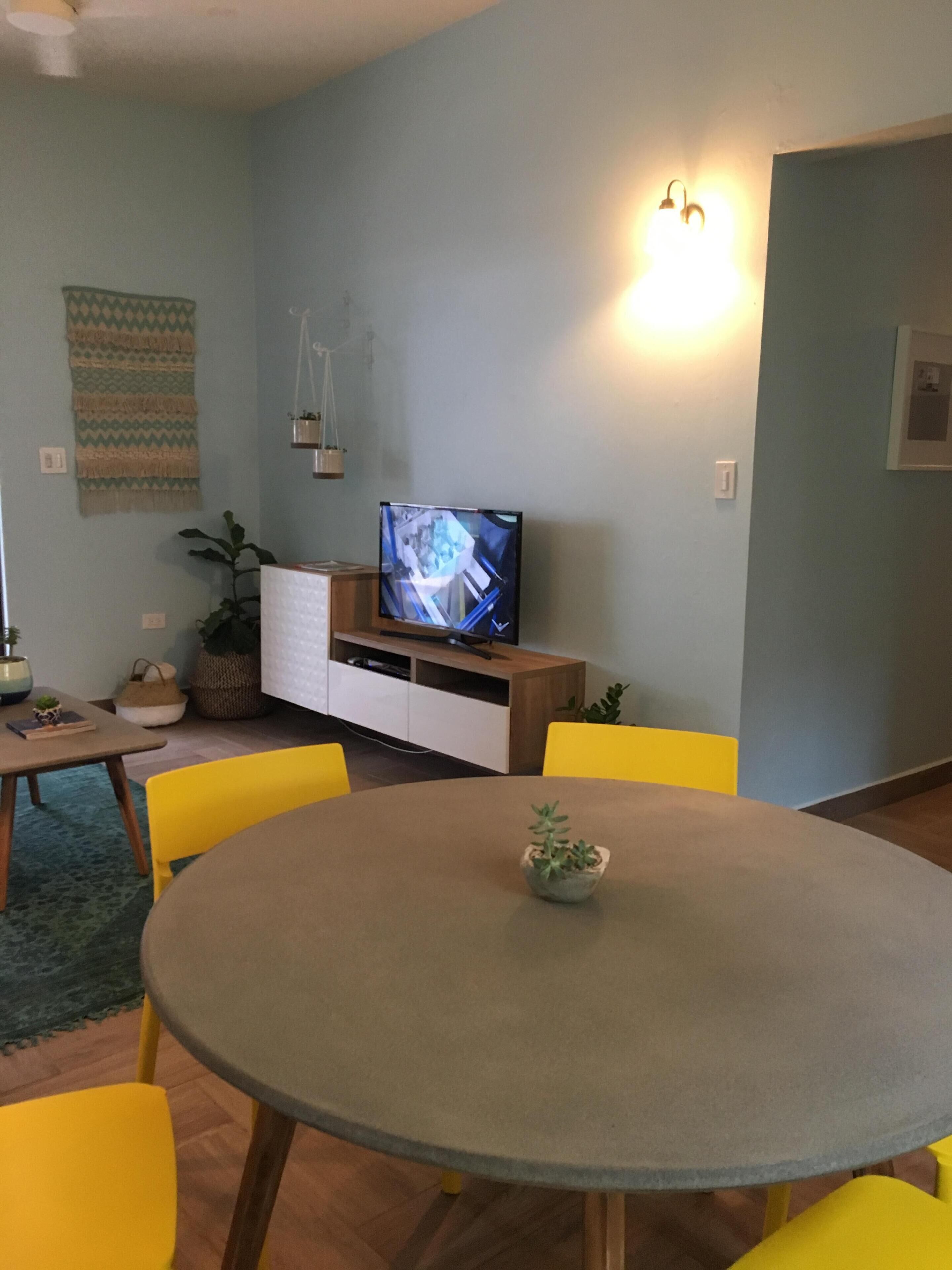 Un comedor con una mesa redonda, sillas amarillas, un televisor sobre un mueble de madera y una lámpara fijada a la pared.