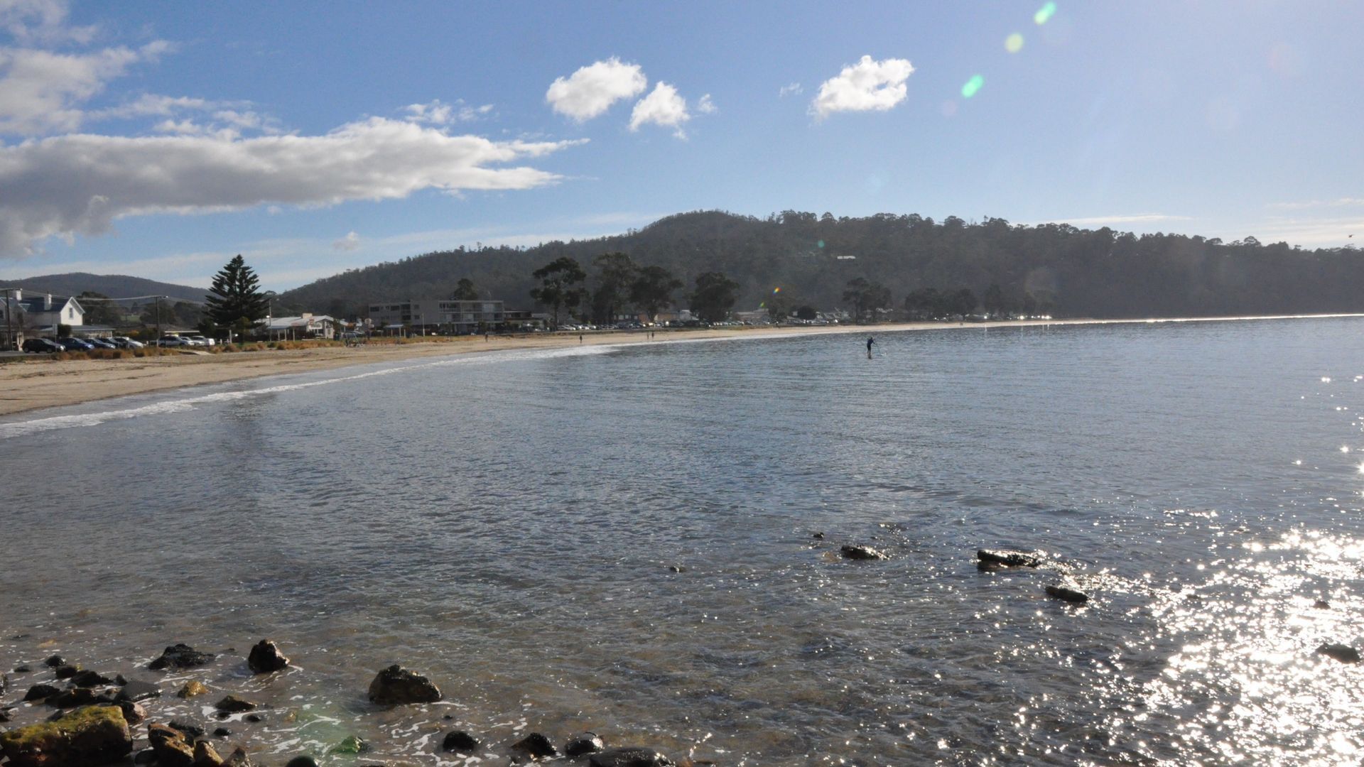Una playa con agua cristalina, rocas en primer plano y una colina boscosa al fondo.