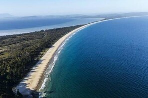 Vista aérea de una playa larga y estrecha, con un océano azul cristalino y un área boscosa junto a la orilla.