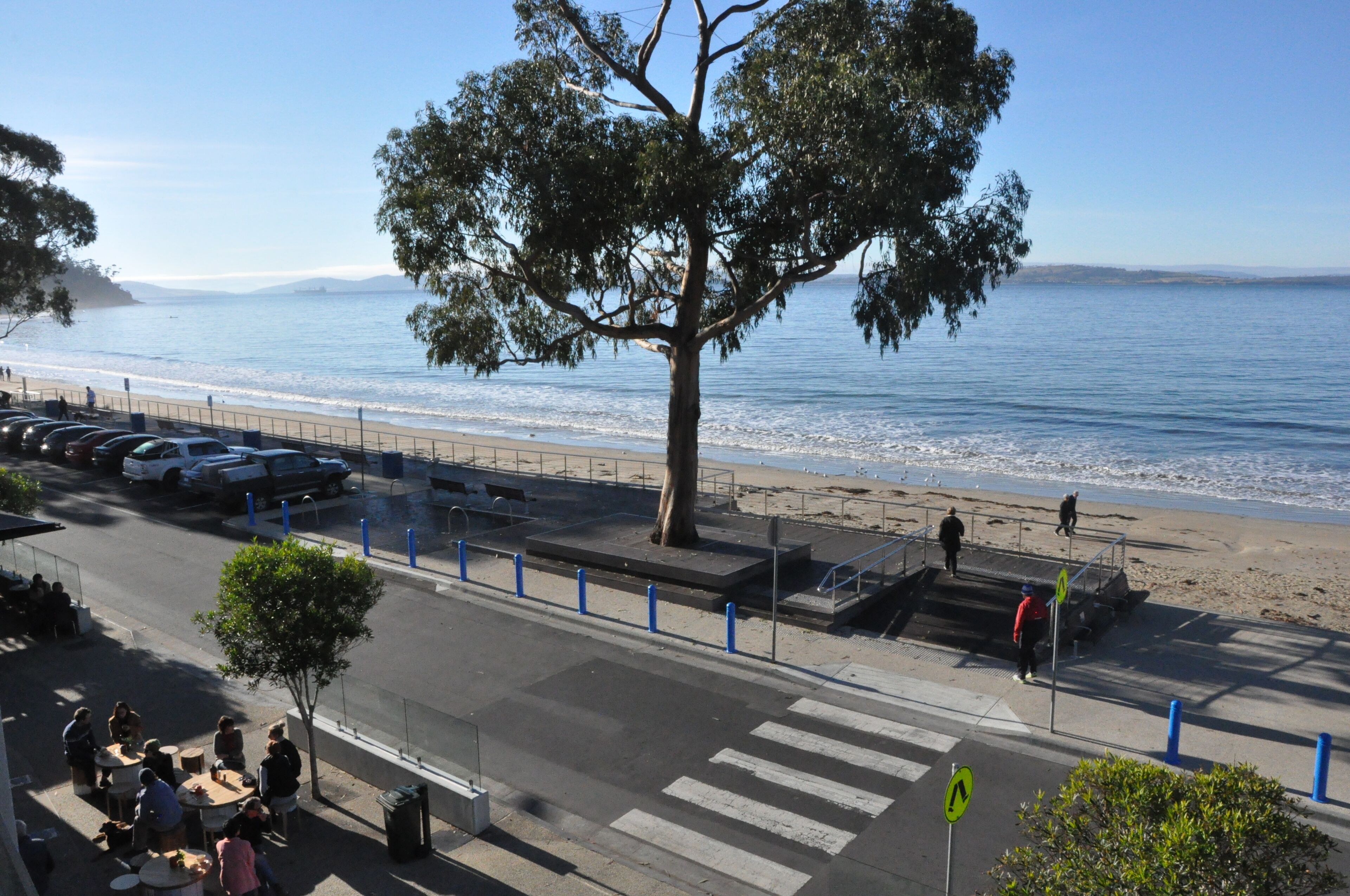 Une route côtière avec un passage piéton, un trottoir bordé d’arbres et une plage en arrière-plan.