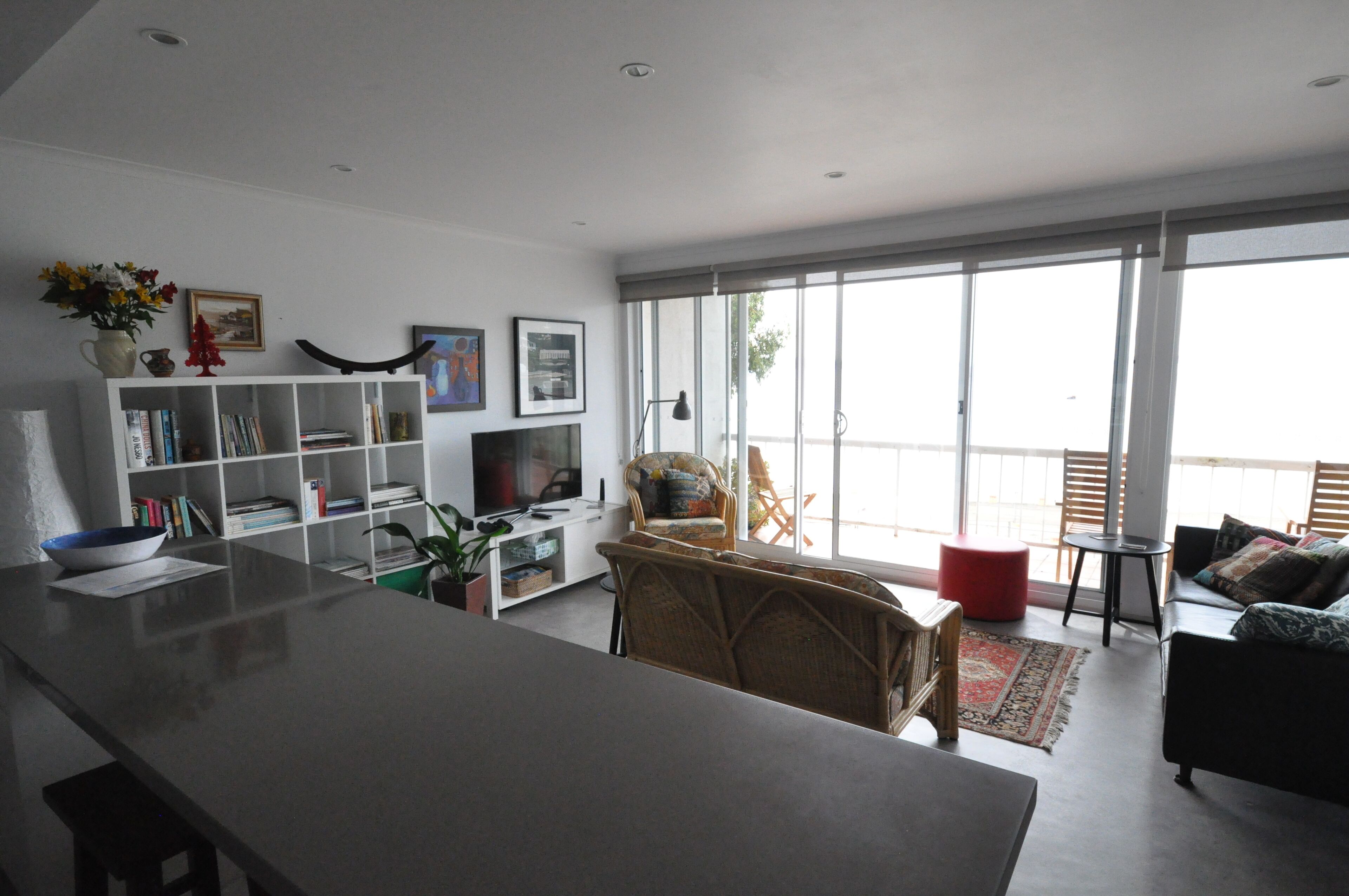 A modern living room with a flat-screen TV, a bookshelf, a dining area with a large table, and a balcony with a view.