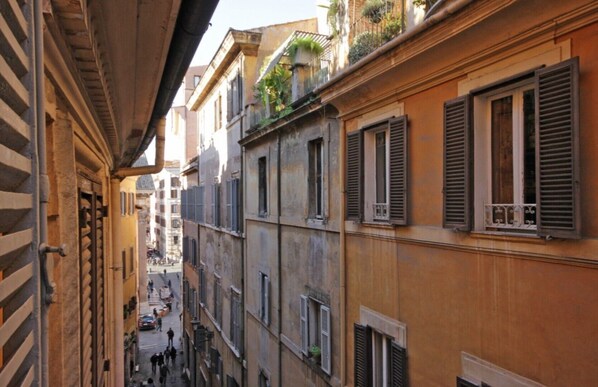 Une rue étroite, bordée de bâtiments des deux côtés, avec des balcons ornés de plantes et de volets.