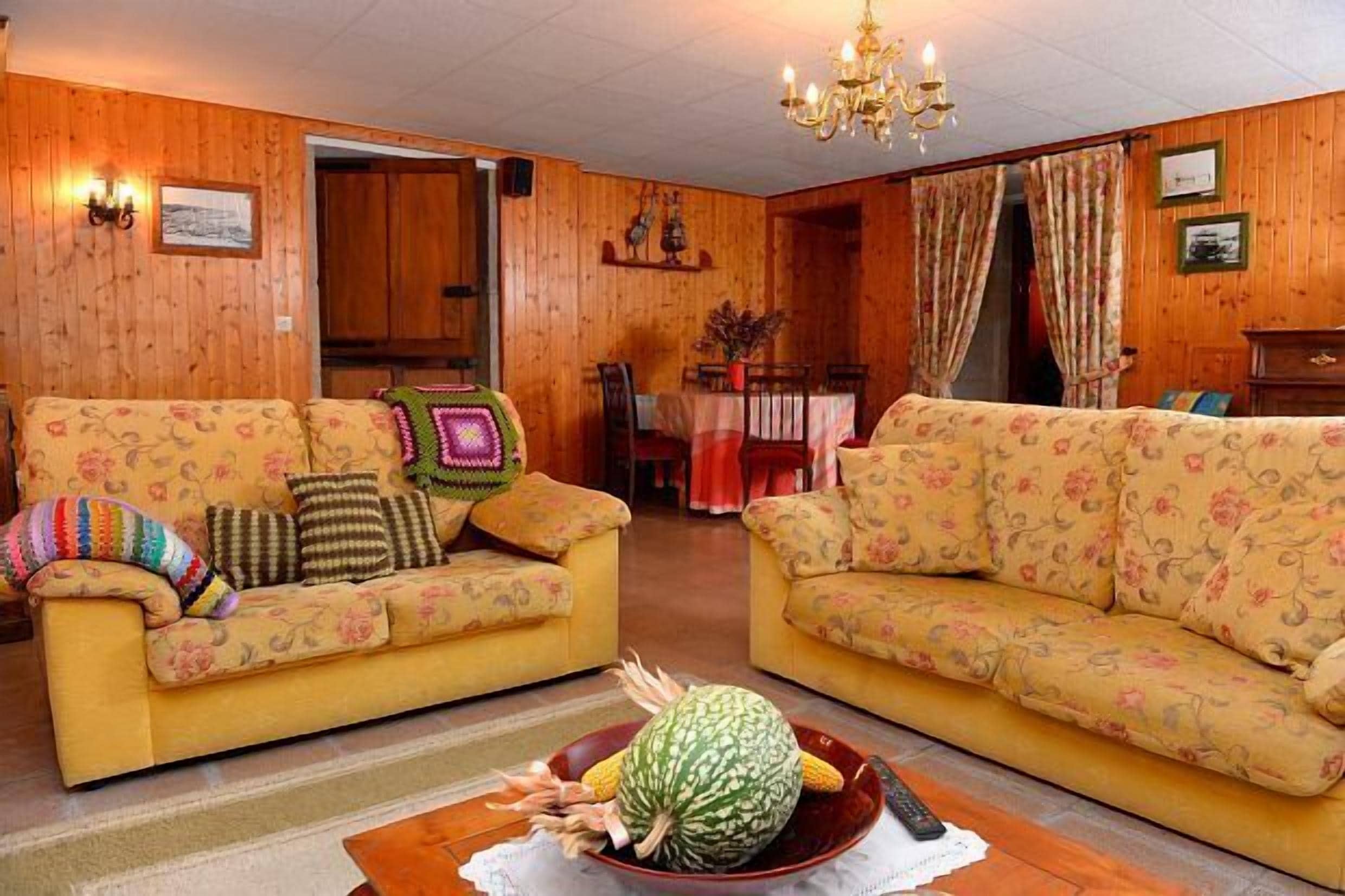Un salón acogedor con dos sofás amarillos con estampado floral, una araña, paredes de madera y un área de comedor al fondo.