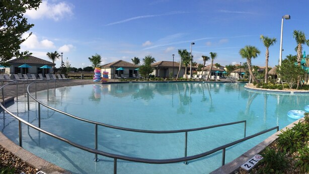 Une grande piscine dont la profondeur maximale est de 60 cm, des chaises longues, des palmiers et un toboggan aquatique.