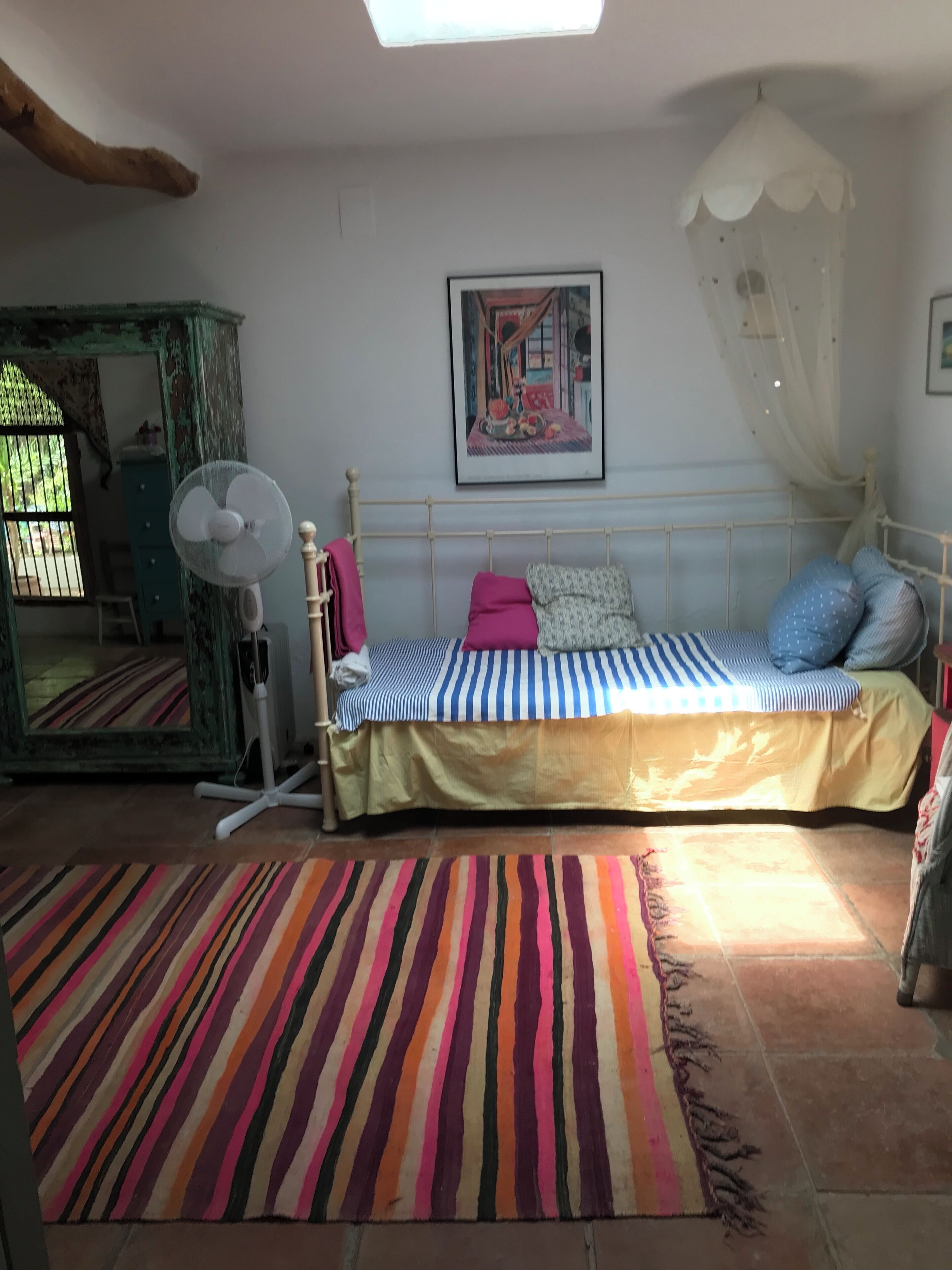 A bedroom with a bed, a standing fan, a colorful striped rug, a painting on the wall, and a window with bars.