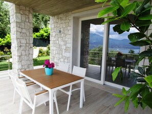 Una zona de estar al aire libre techada con una mesa de madera y sillas blancas, una planta en maceta y vistas a un paisaje montañoso a través de puertas de vidrio.