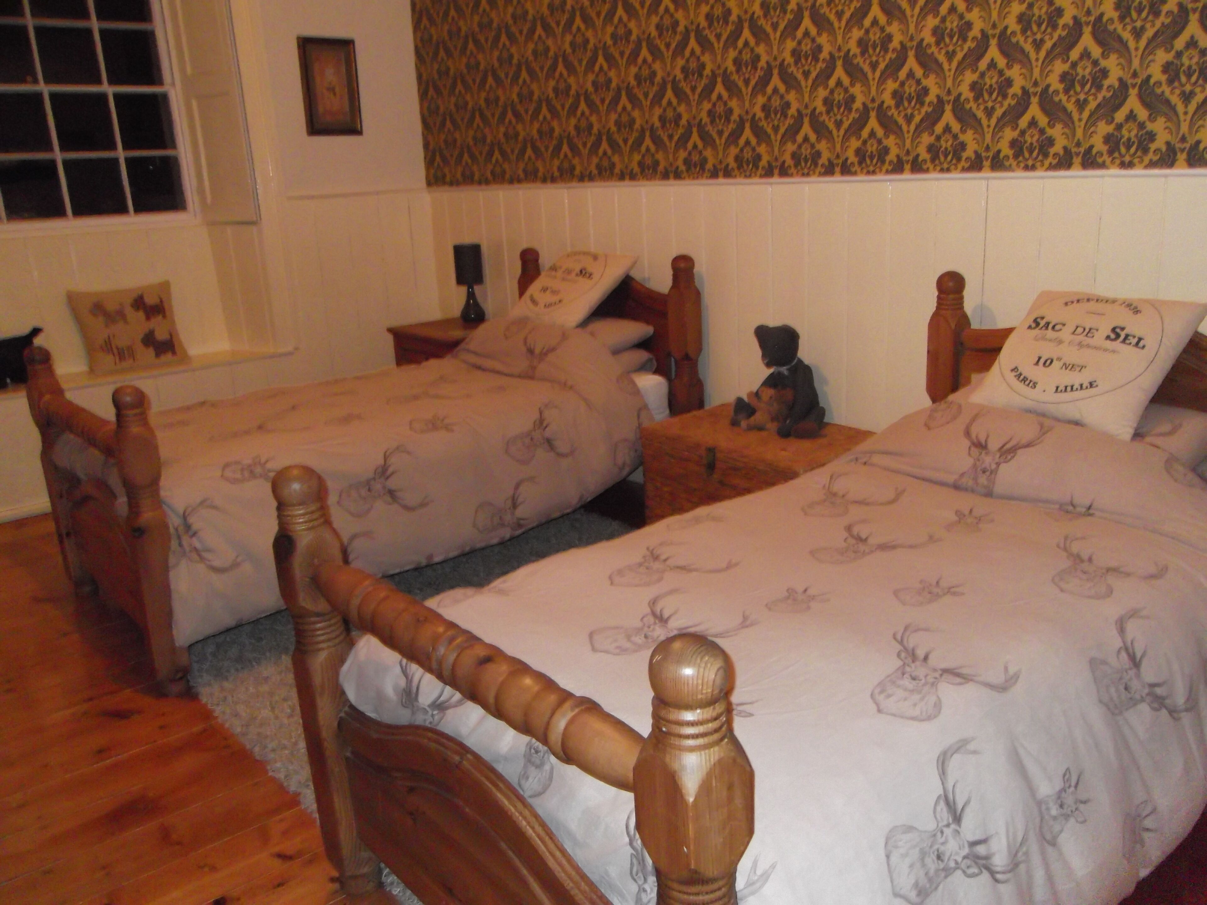 A bedroom with two wooden beds with headboards, a patterned wallpaper, and a small table with a teddy bear.