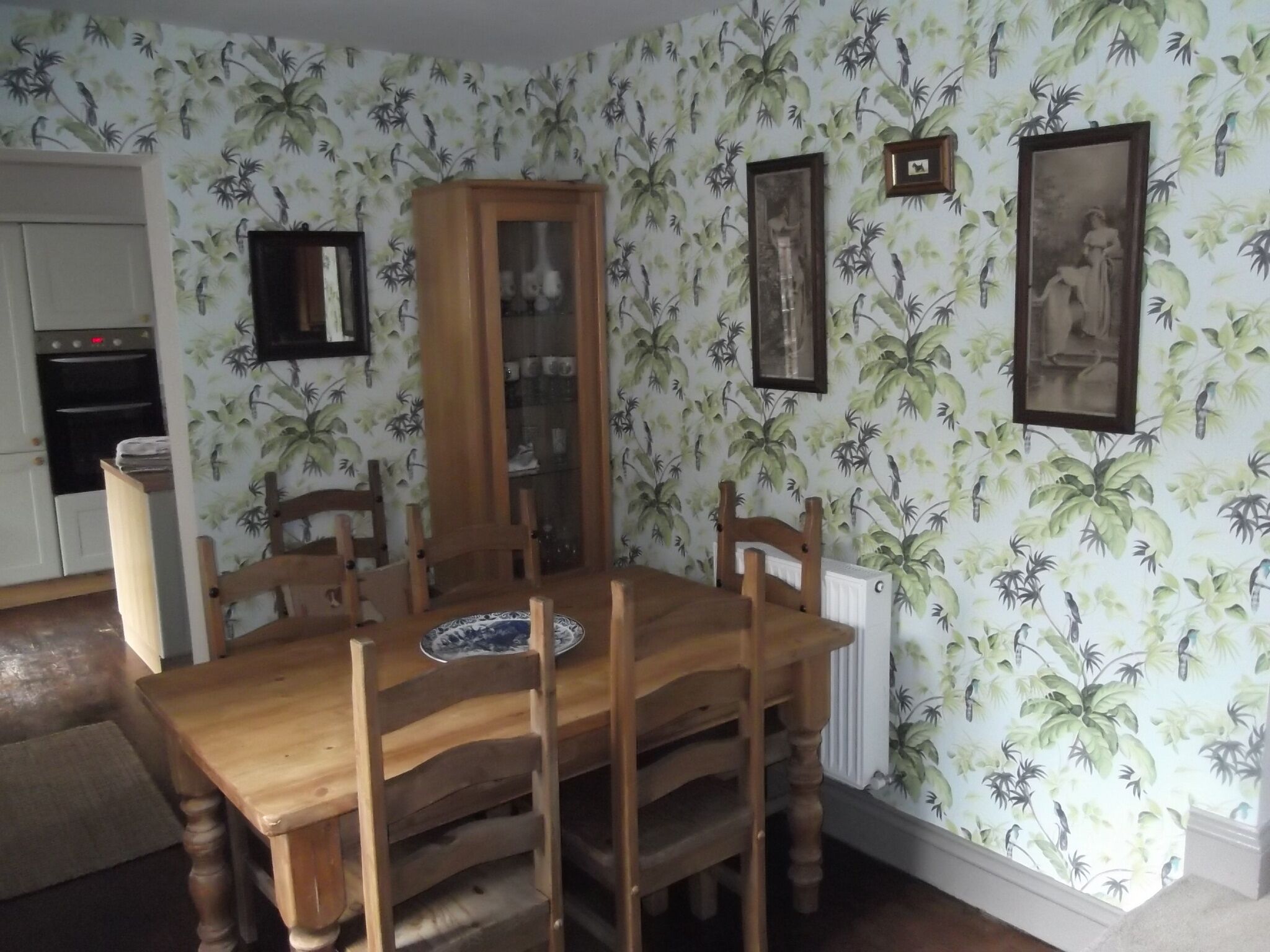 Une salle à manger avec une table et des chaises en bois, un buffet aux portes vitrées et un mur recouvert d’un papier peint aux motifs tropicaux.