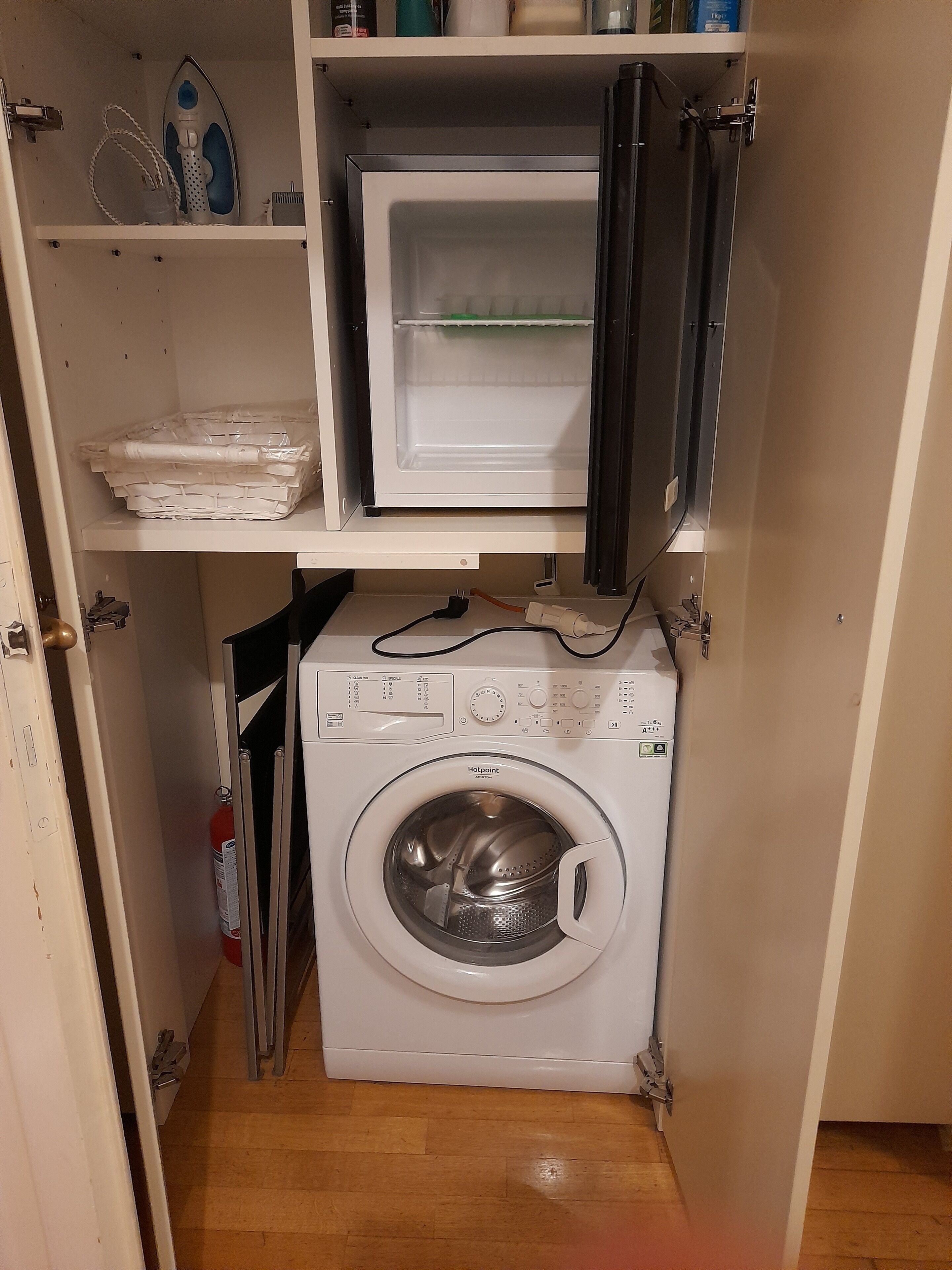 A compact laundry room with a front-loading washing machine, a drying rack, and a shelf above containing a few items.