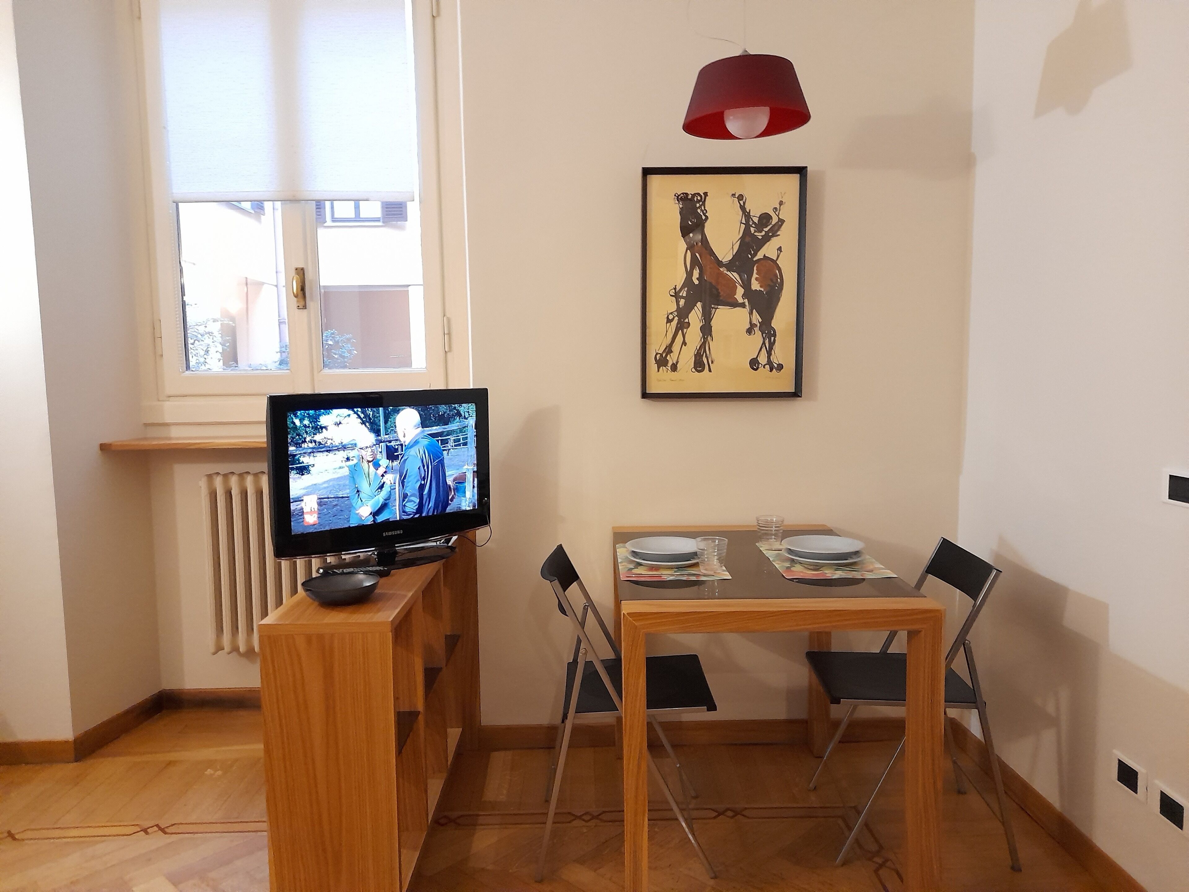 Una habitación con televisión sobre un mueble de madera, una mesa de comedor con sillas, un radiador y una ventana con persianas.