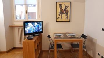 Una habitación con televisión sobre un mueble de madera, una mesa de comedor con sillas, un radiador y una ventana con persianas.