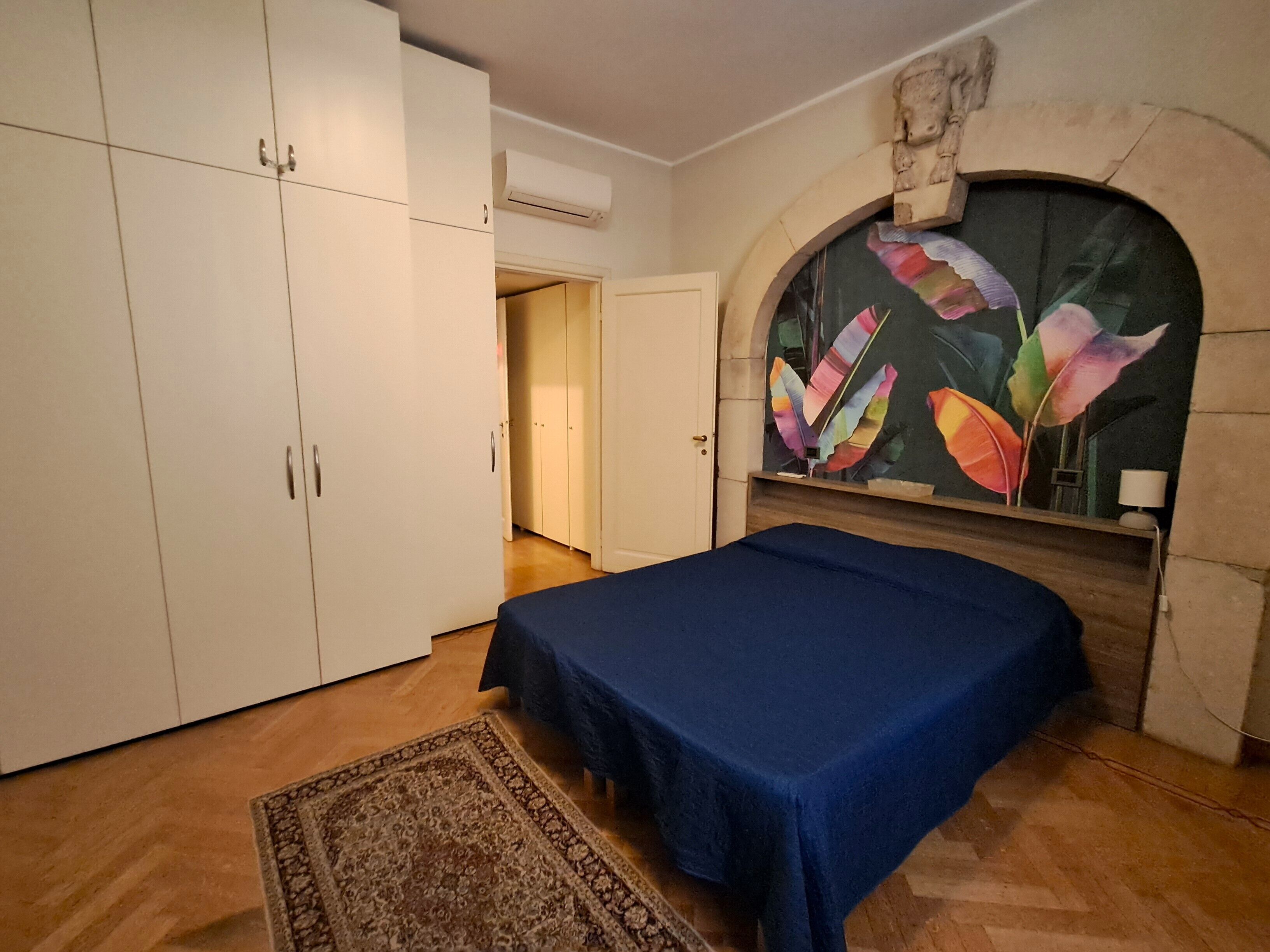 A bedroom with a bed, wardrobe, and a fireplace with a colorful leaf painting.