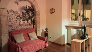 Um quarto acolhedor com um sofá vermelho estampado, um arco de pedra, um mural com vista para a cidade, um radiador de madeira e uma televisão sobre um suporte de madeira.