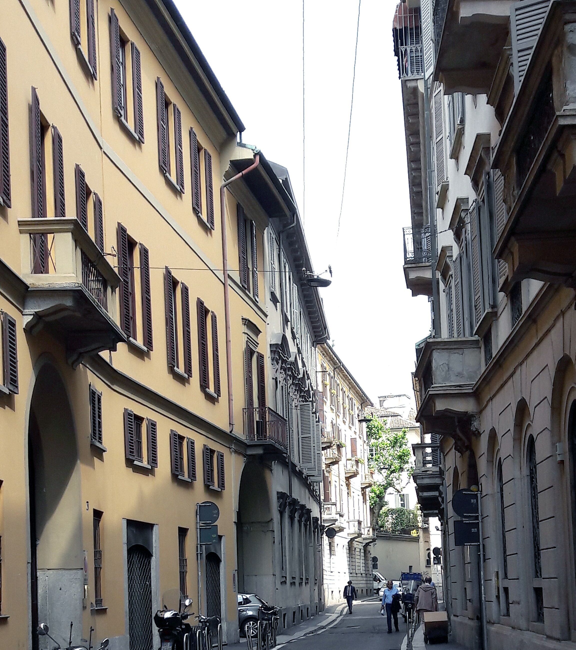 Une rue étroite bordée de bâtiments historiques, de balcons et où quelques véhicules sont stationnés.