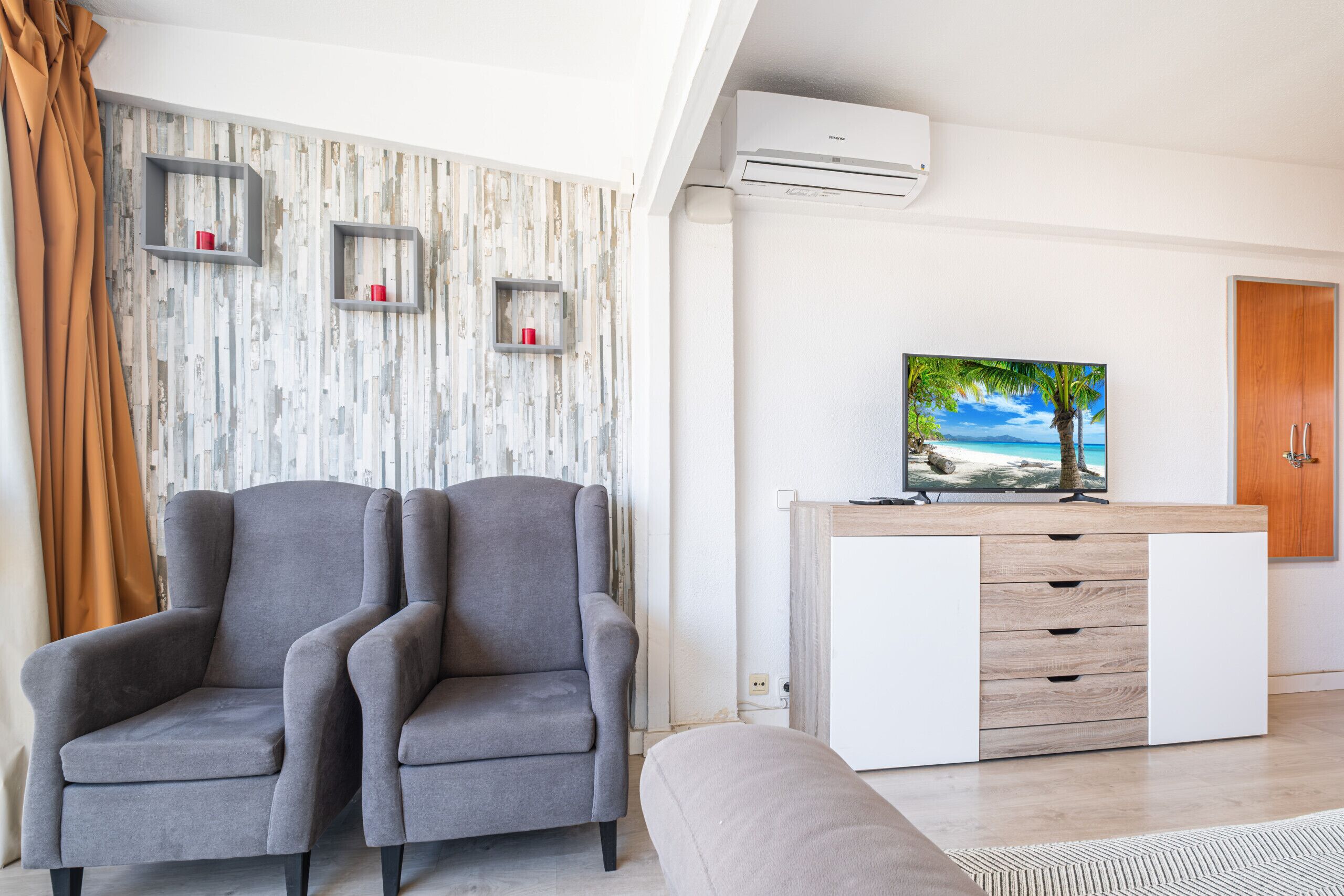 Un salón moderno con dos sillones grises, un mueble blanco con cajones, un televisor de pantalla plana sobre un pedestal de madera y una pared con estampado.