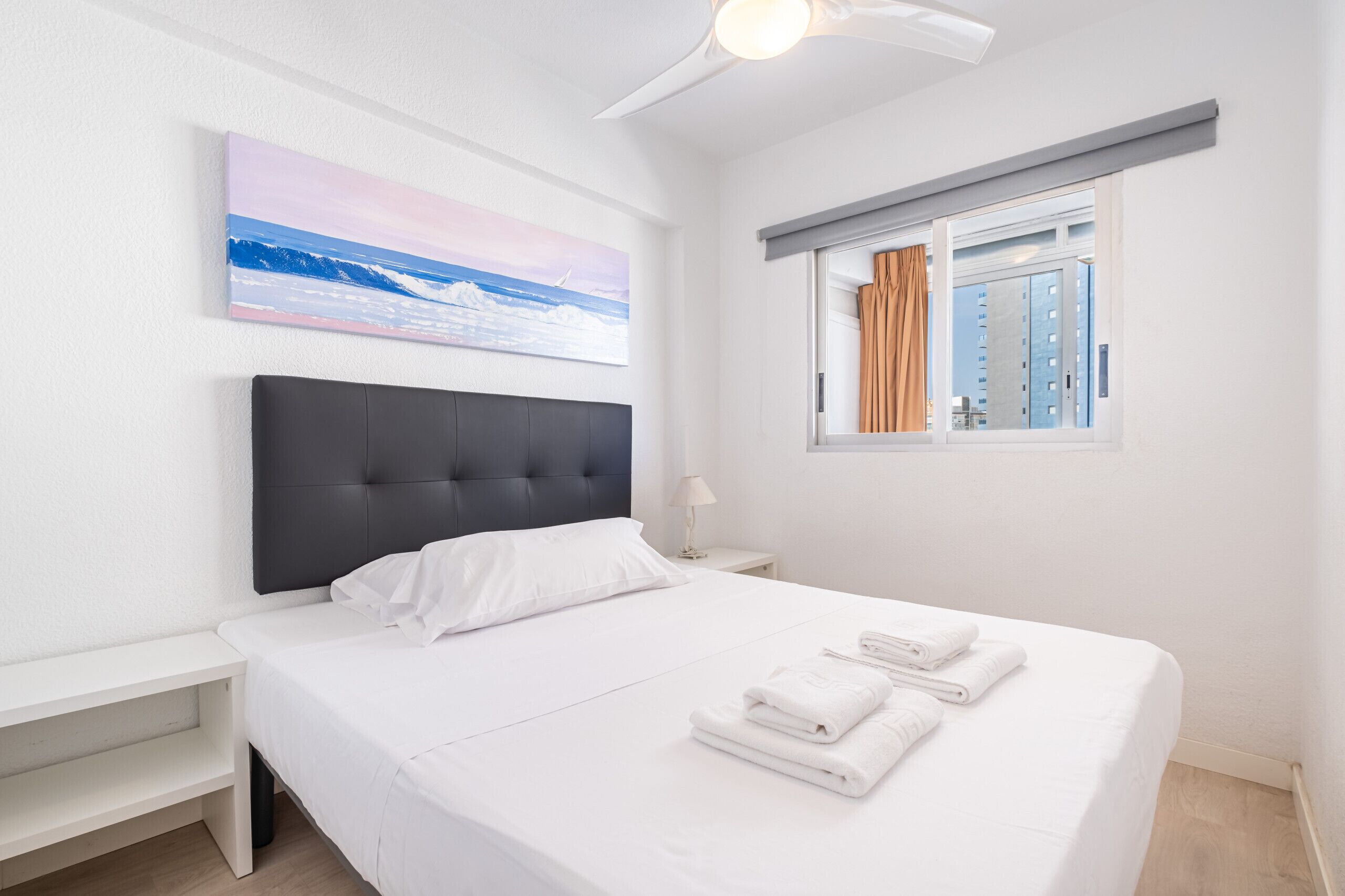 A neatly made bed with white linens and towels, a dark headboard, a bedside table with a lamp, and a window with curtains.