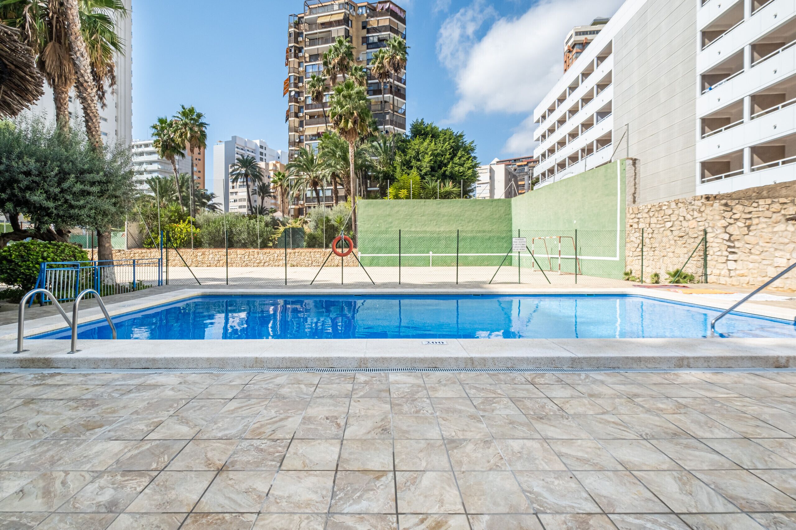 Piscina com deck de pedra, rodeada por edifícios altos e palmeiras.
