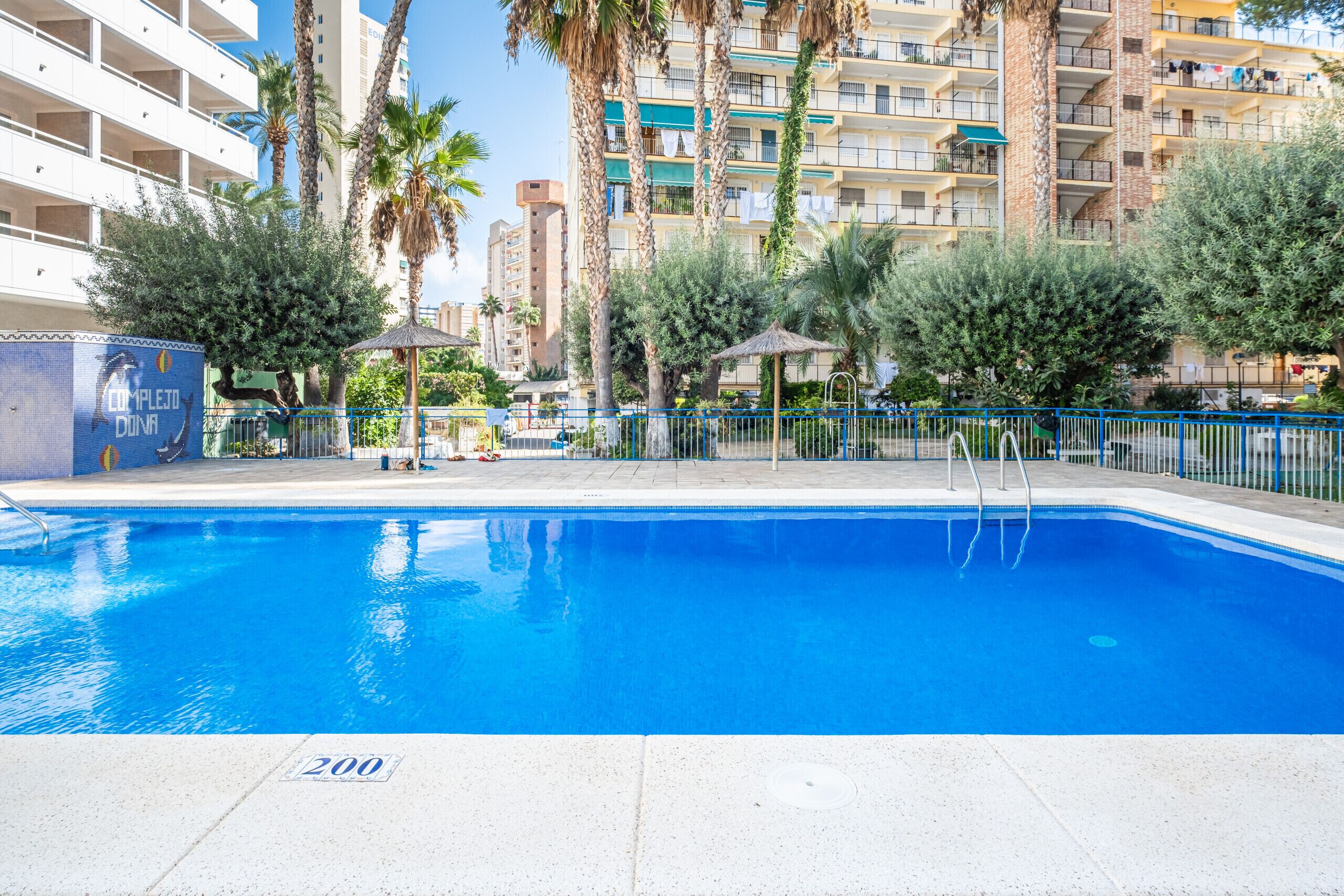 Une piscine à l’eau bleue, entourée d’une terrasse carrelée blanche et d’une clôture bleue.