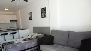 A modern living room with a grey sofa, a bed with a patterned bedspread, and a kitchen area in the background.