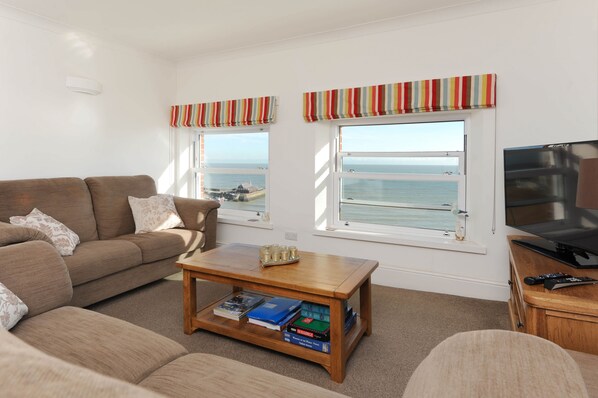A living room with a sofa, a coffee table with books, a TV, and windows with a view of the sea.