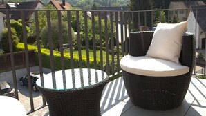 Een balkon met een rieten stoel en een kleine tafel, met uitzicht op een groen landschap met huizen.