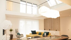 Een moderne woonkamer met een bank, een eettafel en stoelen. Er is een groot raam met gordijnen en een plafondlamp.