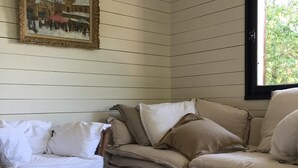 Una habitación con piso de madera, una mesa redonda de madera, un sofá beige con almohadones, un sillón blanco y un cuadro en la pared.