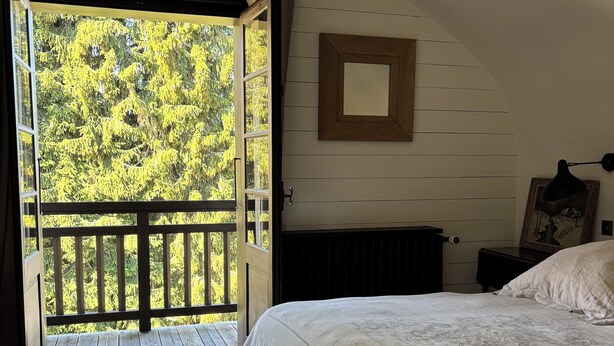 Un dormitorio con una cama grande, piso de madera, vista a árboles desde una puerta abierta y un ventilador de techo.