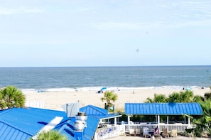 Vista da praia com um edifício de telhado azul, palmeiras e uma praia de areia com pessoas e guarda-sóis.