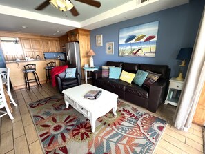 Sala de estar com sofá, mesa de centro e tapete. Há uma cozinha com armários de madeira e geladeira.