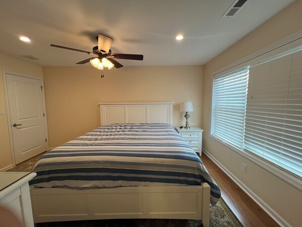 Une chambre à coucher comprenant un lit, une table de chevet, une lampe, un ventilateur de plafond et une fenêtre avec des stores.