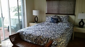 A bedroom with a bed, a chair, a nightstand, a lamp, and a window with blinds. - The Cottage at Villa Indigo 2BR/1.5BA in Exclusive Private Gated Community (Saint Thomas)