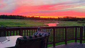 Une terrasse avec une table et des chaises, d’où l’on peut admirer un coucher de soleil sur un champ et l’eau.