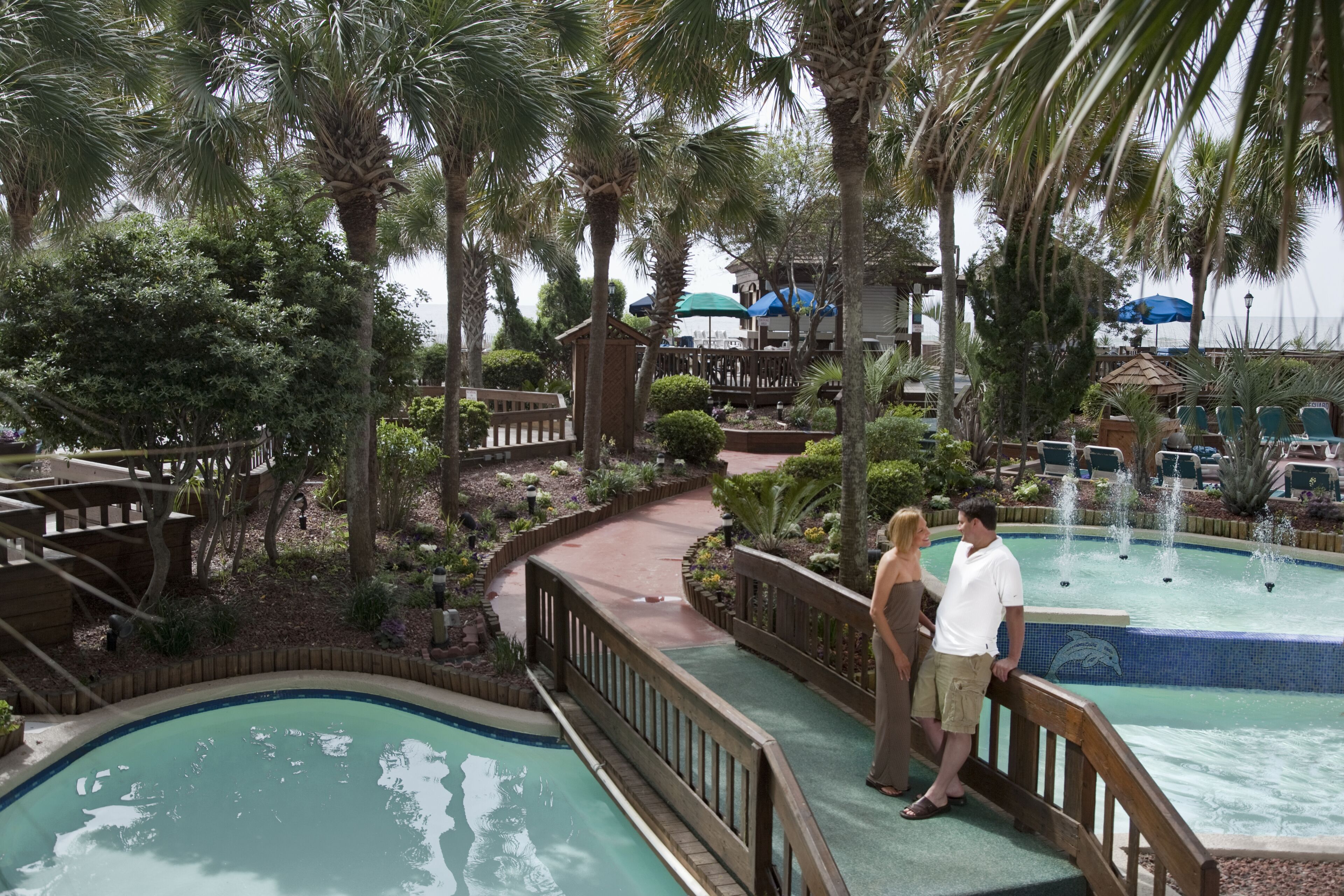 A resort pool area with a wooden deck, palm trees, and a fountain.