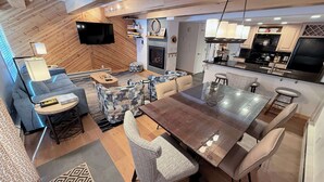 A modern living room with a sofa, armchairs, a coffee table, and a TV. There is a dining area with a table and chairs. The kitchen is visible with a stove and cabinets. - Remodeled Condo with Fireplace - In unit Washer/Dryer (Vail)