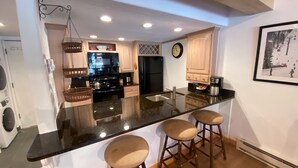 A modern kitchen with a black granite countertop, stainless steel appliances, and wooden bar stools. - Remodeled Condo with Fireplace - In unit Washer/Dryer (Vail)