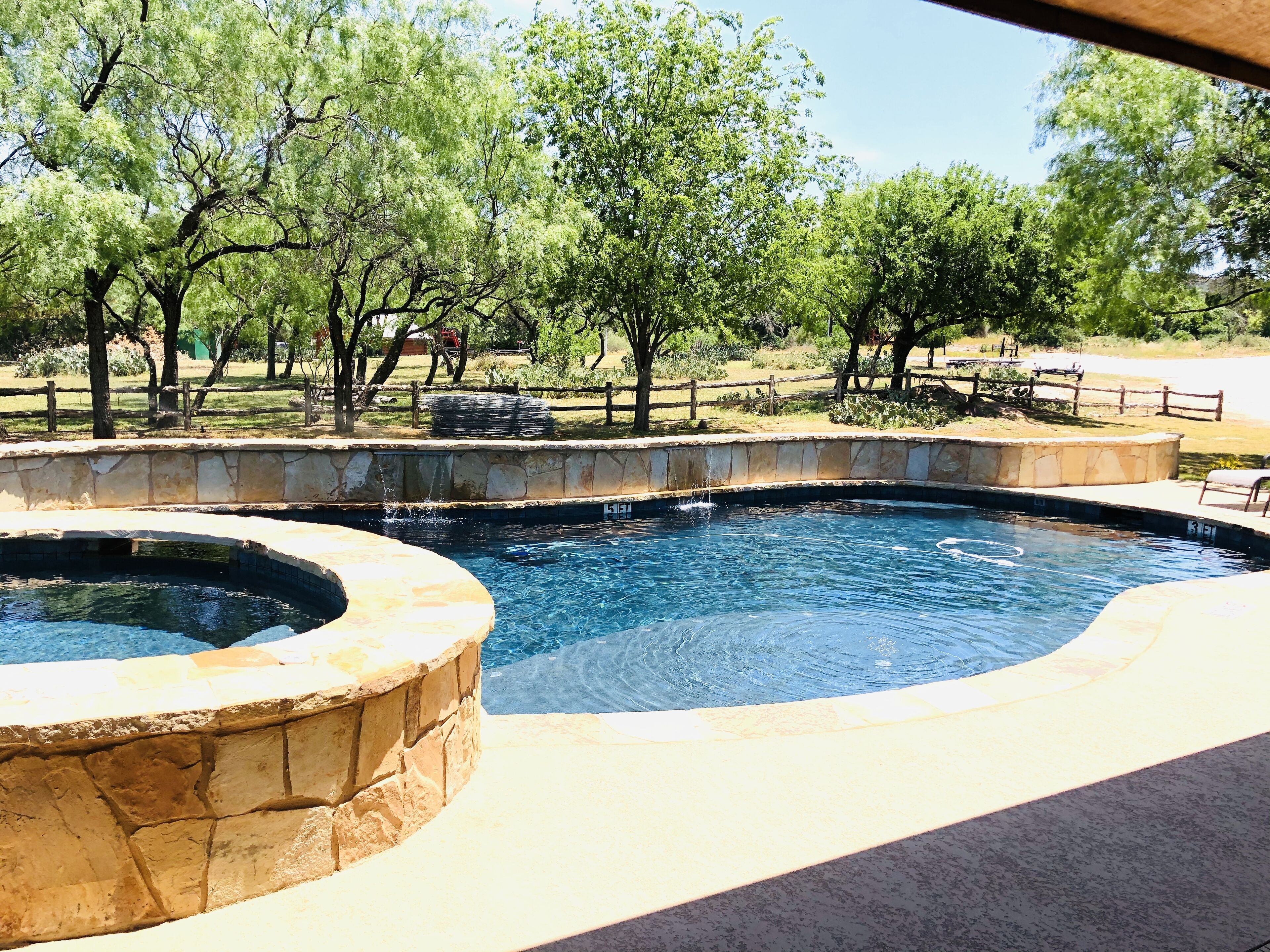 A swimming pool with a stone border and a circular hot tub, surrounded by trees and a wooden fence.