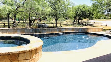 Una piscina con borde de piedra y un jacuzzi circular, rodeada de árboles y una valla de madera.
