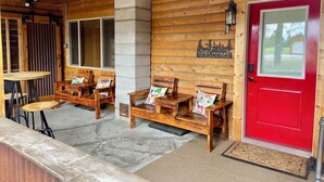 Una cabaña de madera con una puerta roja, bancos de madera con cojines de colores y una pequeña mesa.