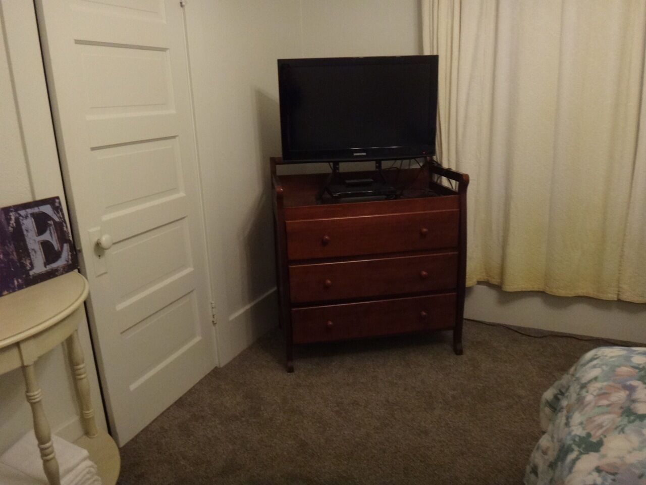 A room with a wooden dresser, a television on top, a door, and a bed with a floral bedspread.