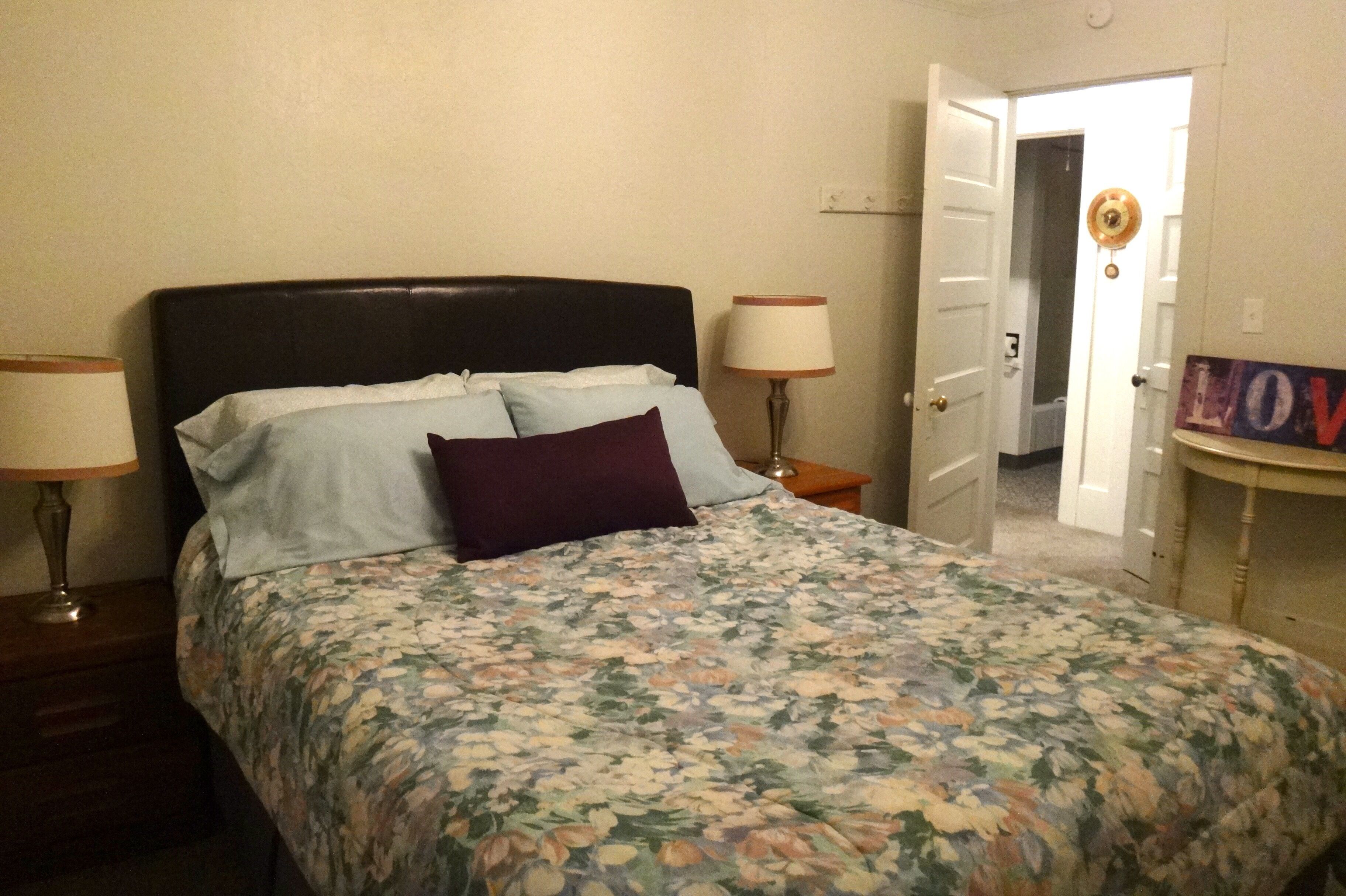 A bedroom with a bed, two bedside tables, a lamp, and a door leading to another room.