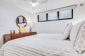 A bedroom with a bed, a ceiling fan, a window with blinds, a bedside table with a lamp, and a mirror. - Modern Ground Floor Retreat - Kihei Surfside #110 - Perfect Oceanfront Grounds! (Kihei, Maui)