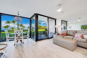A spacious living area with a sliding glass door leading to a balcony with a view of palm trees and a clear sky. - Modern Ground Floor Retreat - Kihei Surfside #110 - Perfect Oceanfront Grounds! (Kihei, Maui)