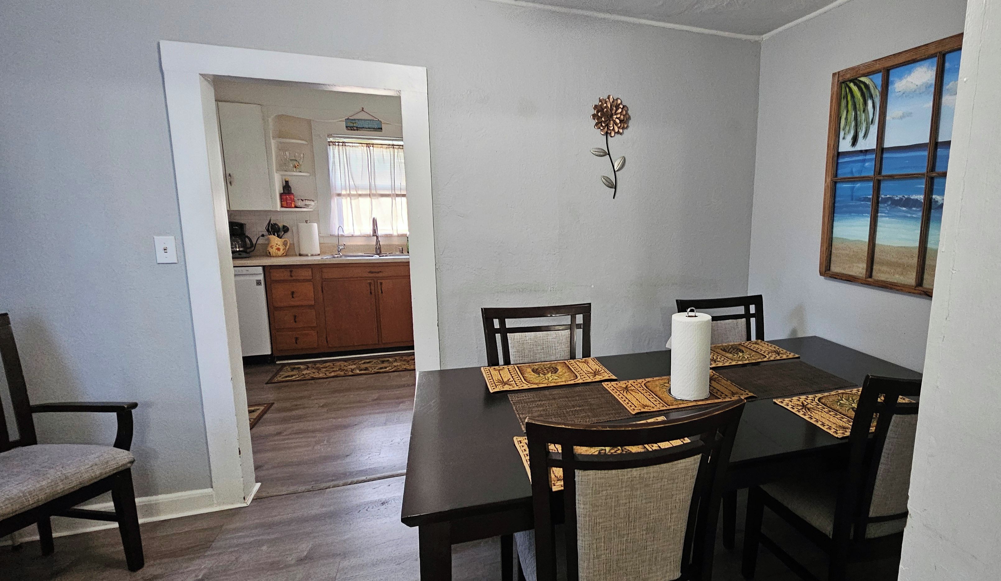 A dining area with a table set for four, a kitchen in the background, and a beach-themed painting on the wall.