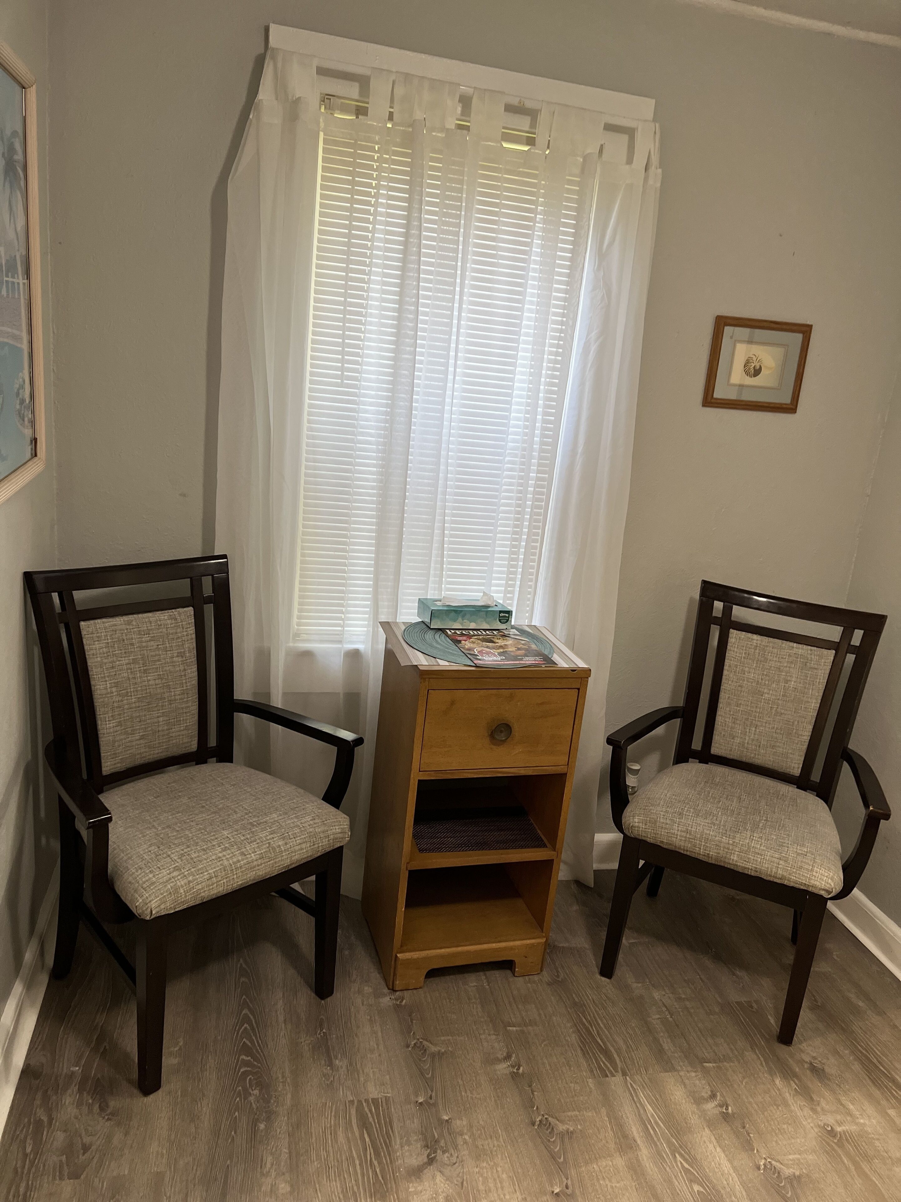 Una habitación con dos sillas de madera con asientos acolchados, una mesita de noche de madera con cajón y una ventana con cortinas blancas.