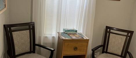Una habitación con dos sillas de madera con asientos acolchados, una mesita de noche de madera con cajón y una ventana con cortinas blancas.