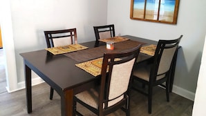 Ein Essbereich mit einem dunklen Holztisch und vier Stühlen, jeweils mit gewebtem Sitzkissen. Der Tisch ist mit gewebten Tischsets mit einem Ananas-Muster geschmückt. An der Wand über dem Tisch hängt ein gerahmtes Bild einer Strandansicht.