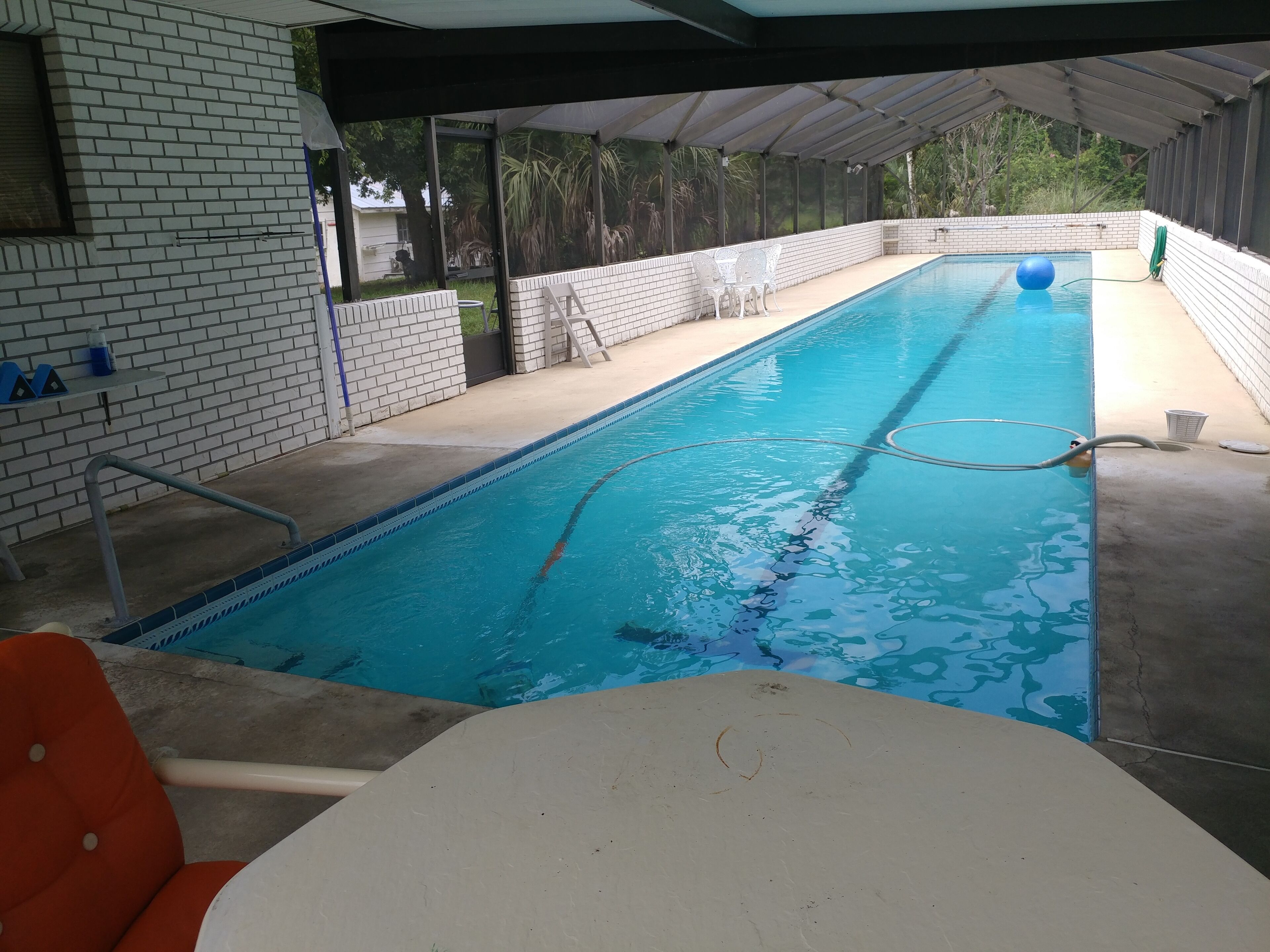 A swimming pool with a tiled deck, a diving board, and a covered area with chairs.