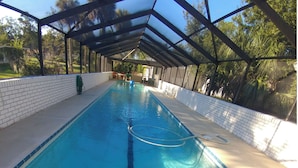 Una piscina larga y rectangular con agua azul cristalina, rodeada por una zona cubierta con pared de ladrillo y estructura de techo metálico.
