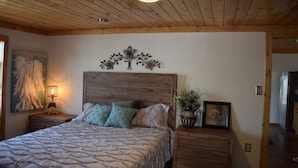 A bedroom with a wooden ceiling, a bed with a quilted blanket, a nightstand with a lamp, and a wall-mounted decorative piece. - Allure 1900 sq waterfront home with country elegance spectacular views (Eastland)